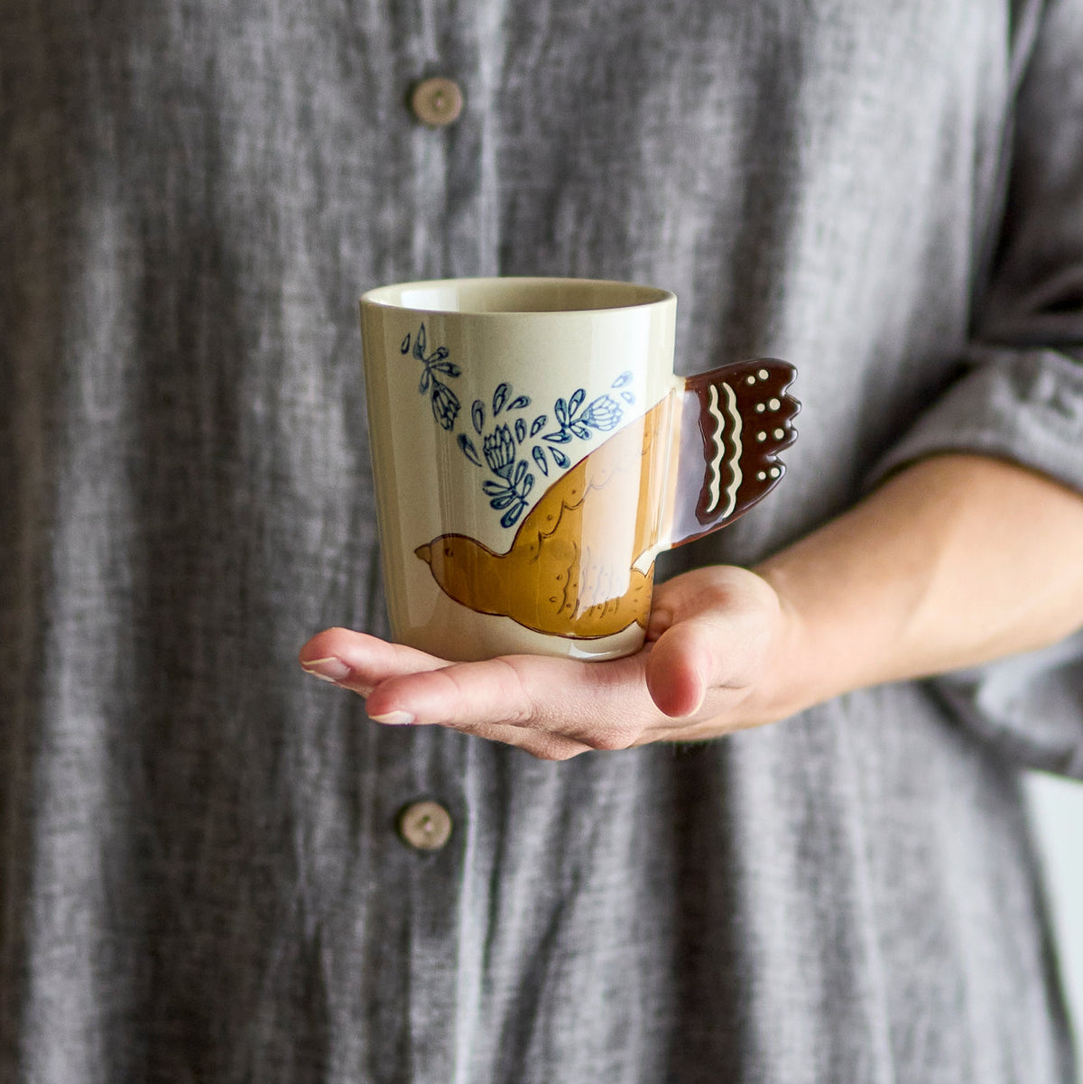 Tasse mit handbemaltem Vogelmotiv und Vogelschwanz als Henkel auf einer Hand.