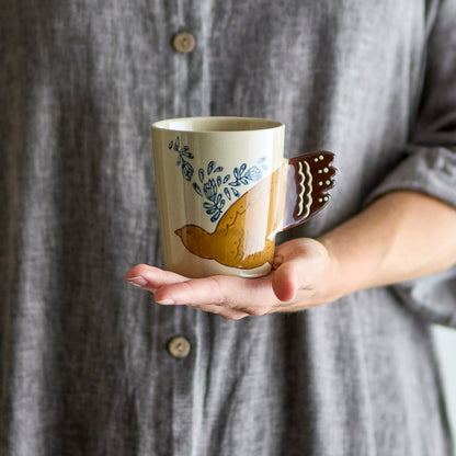 Tasse mit handbemaltem Vogelmotiv und Vogelschwanz als Henkel auf einer Hand.