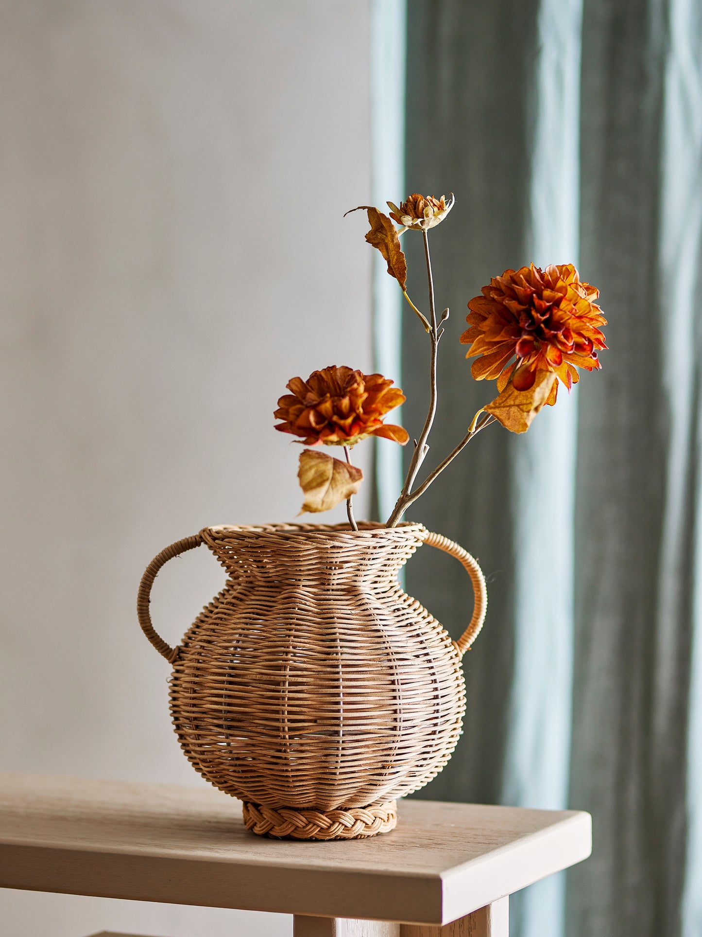 Die runde Rattan-Vase im Boho-Stil dekoriert mit orangen Trockenblumen auf einem hellen Holztisch.
