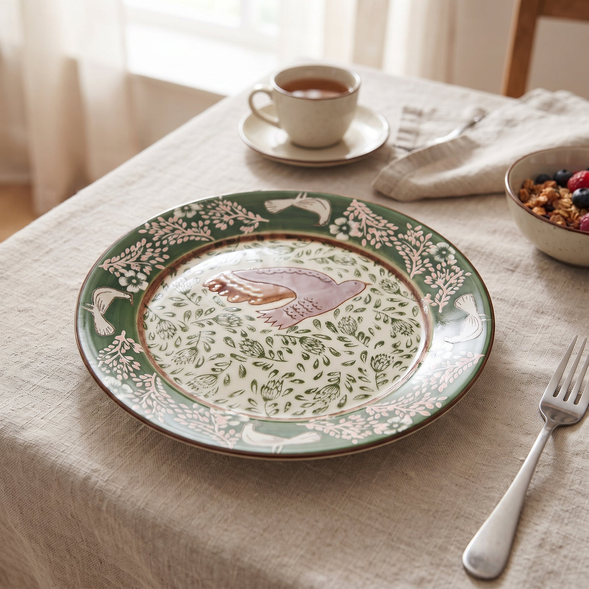 Teller Hezha in grün mit Vogel und Blüten-Muster auf einem Frühstückstisch mit einer hellen Leinen-Tischdecke.