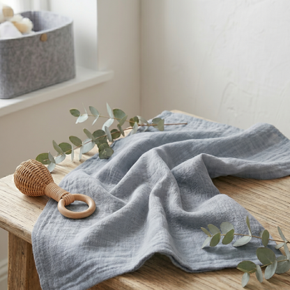Die minimalistische Musselin-Decke als sanftes und funktionales Accessoire gemütlich drapiert auf einem Holztisch mit Rassel und Eukalyptuszweig.