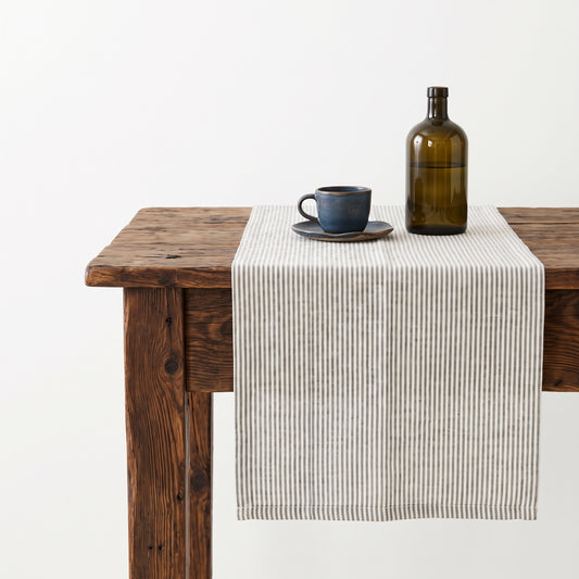 Dunkler Holztisch mit dem grau-weiß gestreiften Leinen-Tischläufer, Steingut-Tasse und grüne Altglas-Flasche.