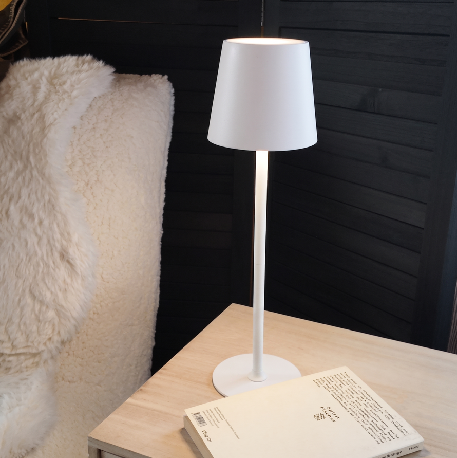 Die gedimmte Akku-Tischleuchte als gemütliche, warme Lichtquelle auf einem hölzernen Nachttisch mit Buch.