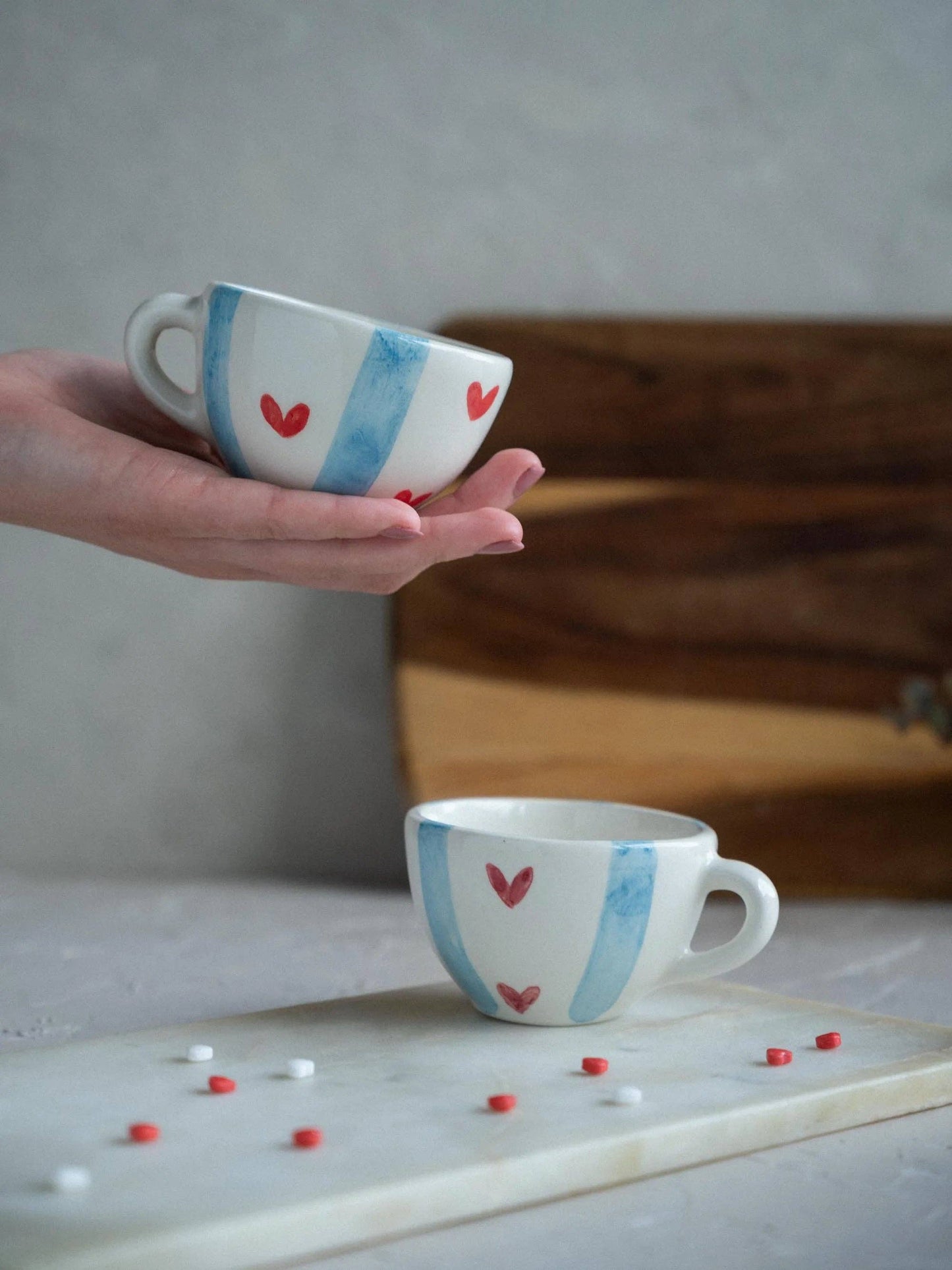 Tassenset Love Stripes (2er Set) mit blauen Streifen und roten Herzen in der Gesamtansicht auf einer Hand und auf einer Holzplatte.