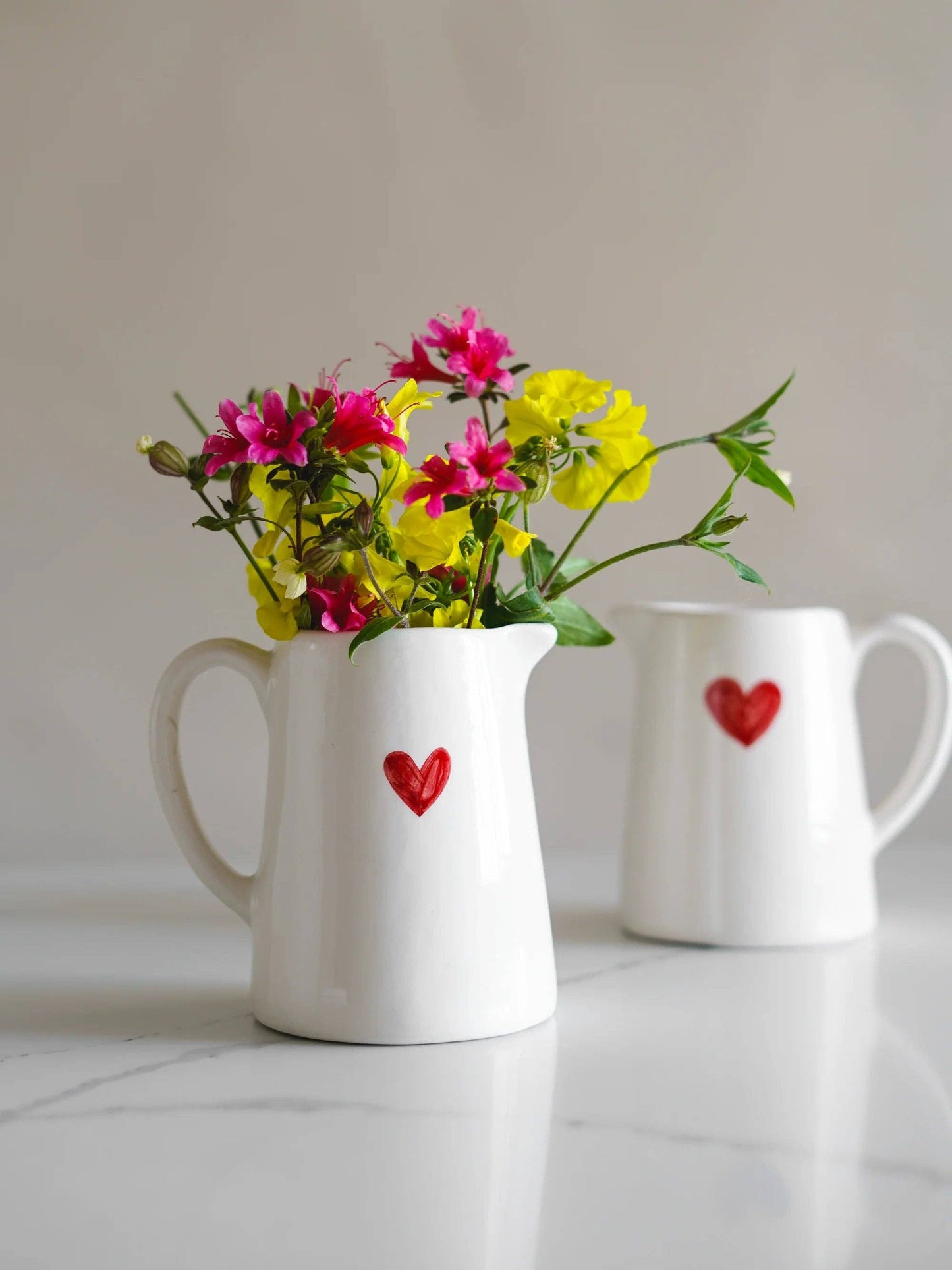 Rustikaler Holztisch mit dem weißen Herz-Milchkrug als Blumenvase mit Wiesenblumen auf einem hellen Marmortisch.