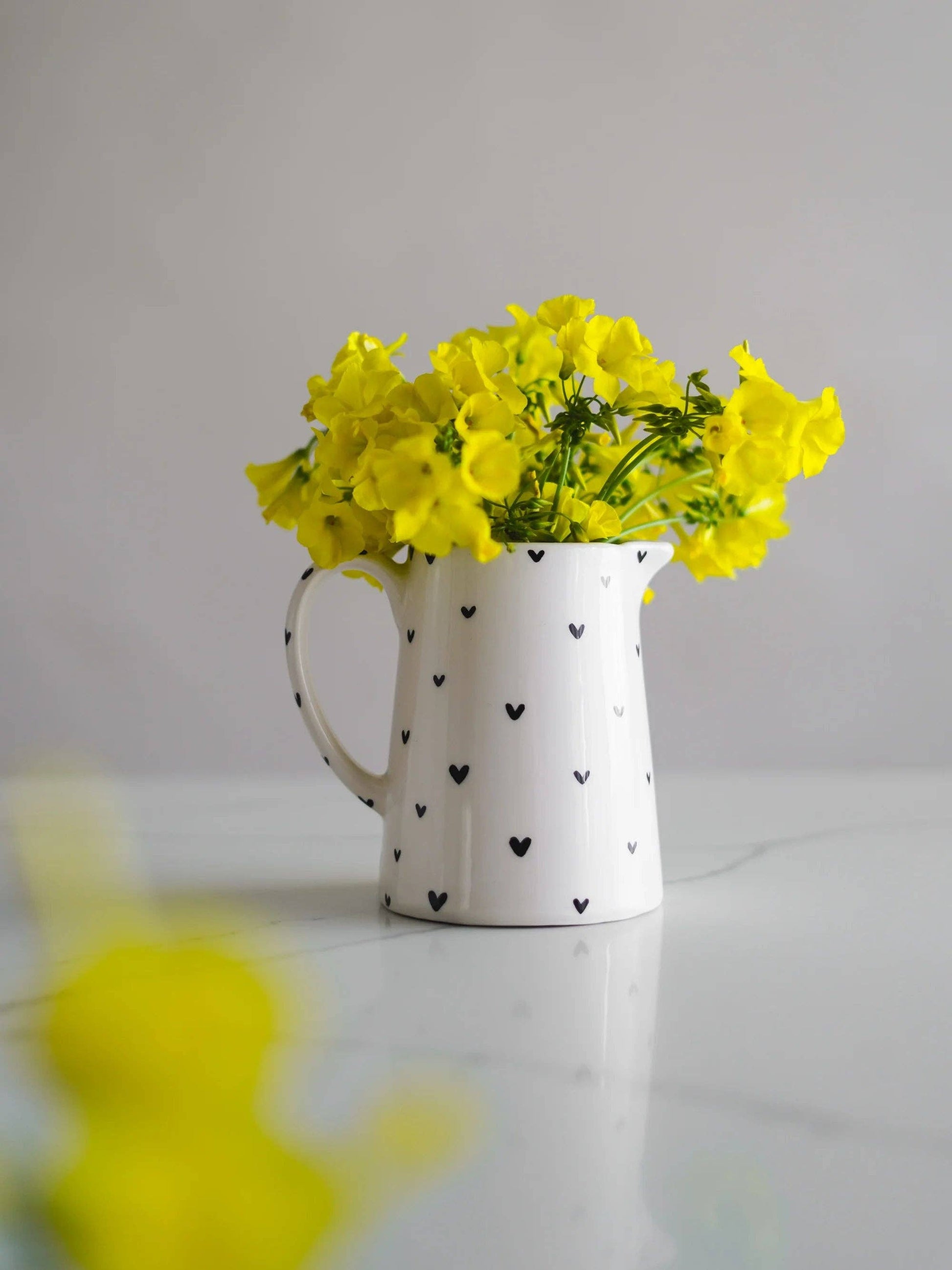 Handgefertigter weißer Keramik-Milchkrug aus Steingut mit schwarzen Herzen als Vase mit gelben Blumen auf einem Marmortisch.