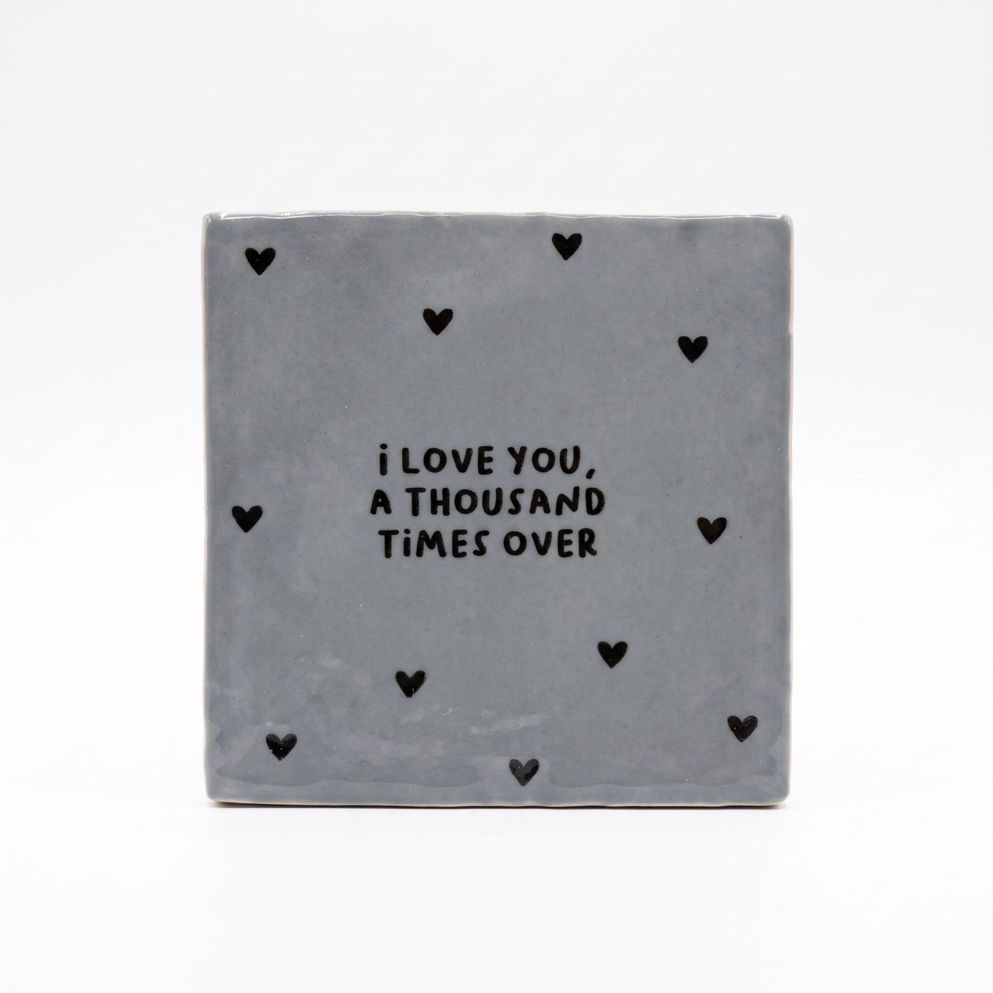 Blau graue Dekokachel mit schwarzer Aufschrift I love you a thousand times over mit vielen kleinen schwarzen Herzen in der Gesamtansicht auf weißem Hintergrund.
