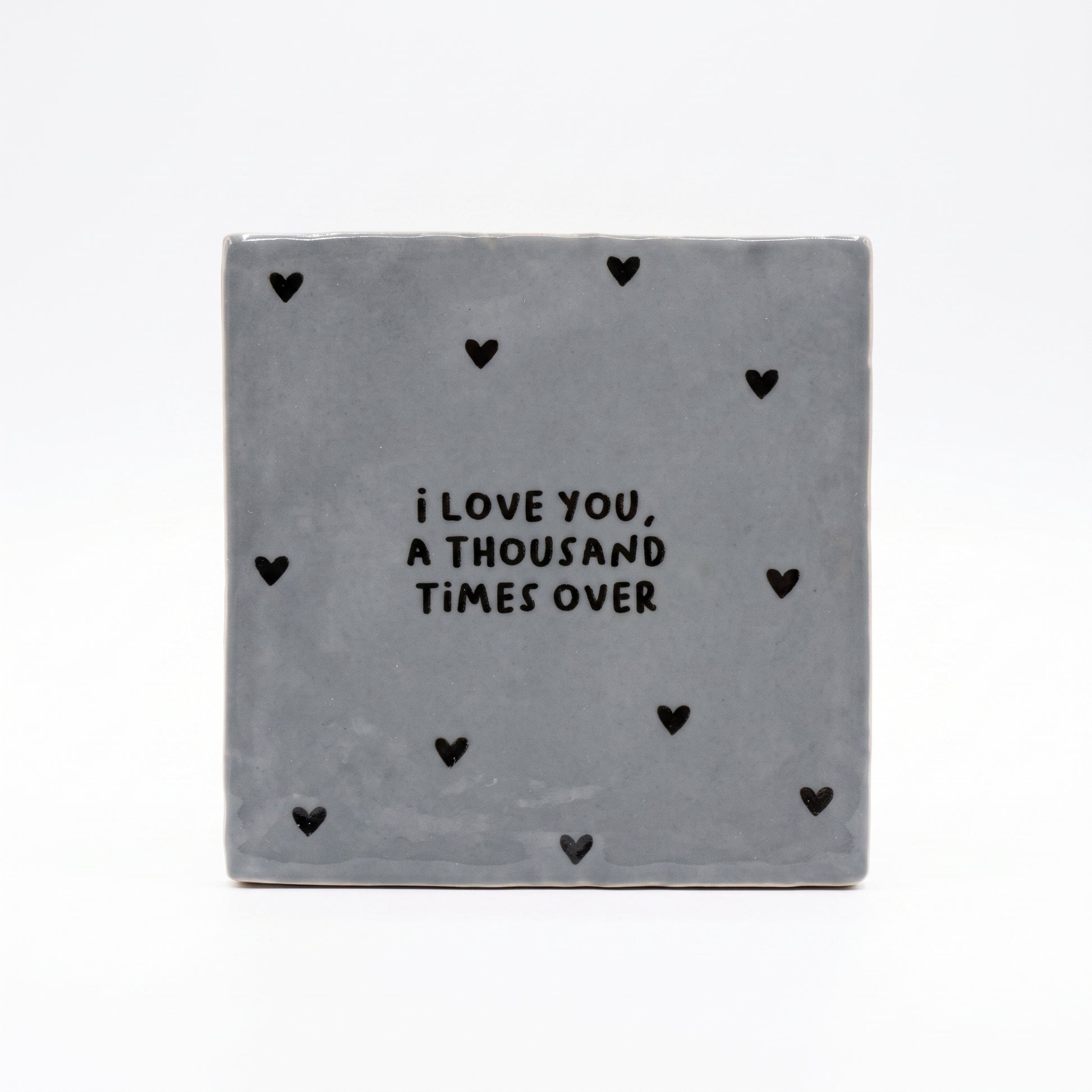 Blau graue Dekokachel mit schwarzer Aufschrift I love you a thousand times over mit vielen kleinen schwarzen Herzen in der Gesamtansicht auf weißem Hintergrund.