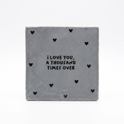 Blau graue Dekokachel mit schwarzer Aufschrift I love you a thousand times over mit vielen kleinen schwarzen Herzen in der Gesamtansicht auf weißem Hintergrund.